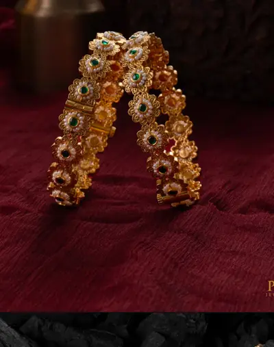 A pair of floral gold bangles set with pearls, standing on maroon cloth.