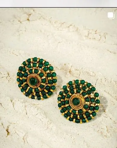Round gold studs ringed with green beads, laid on pale sand.